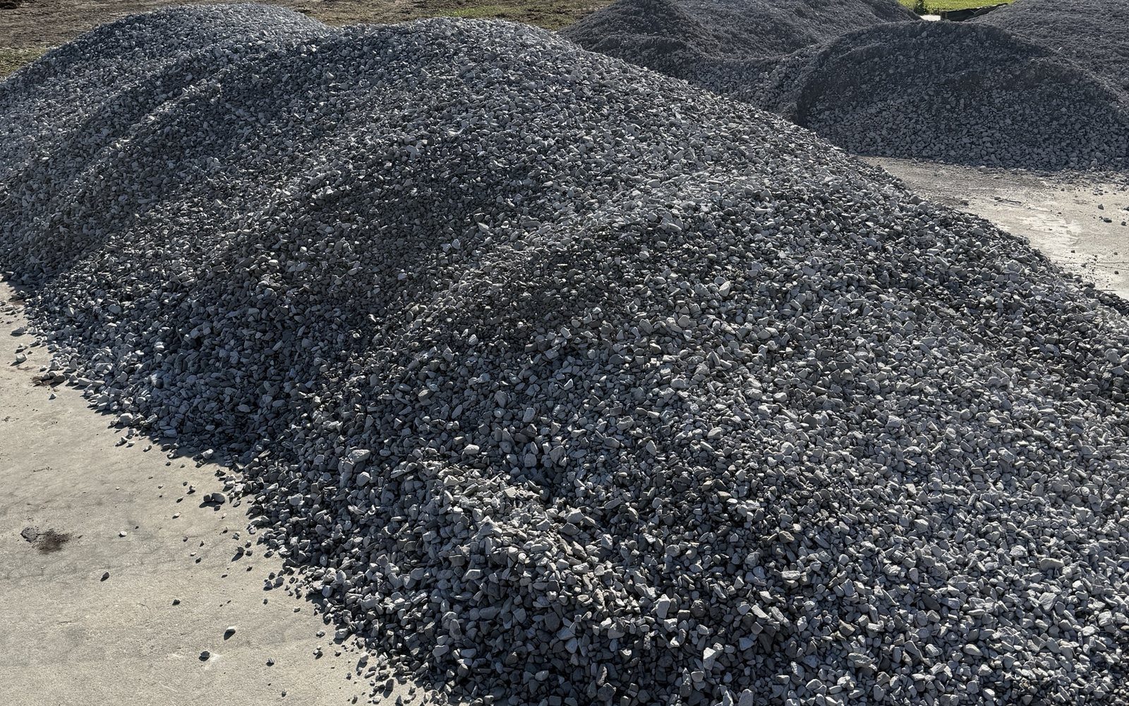 Several truckloads of crushed gravel dumped in long piles on a concrete slab, ready to be spread