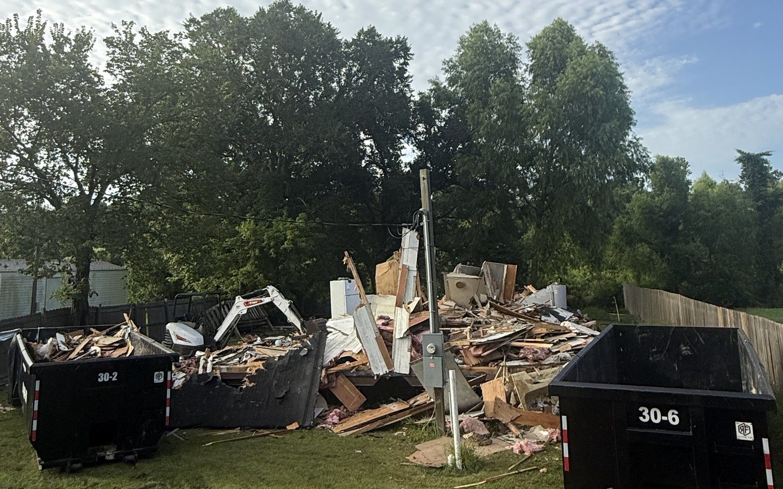 A torn-down house in a pile of lumber and siding, with a mini excavator loading it into two 30 yard roll-off dumpsters