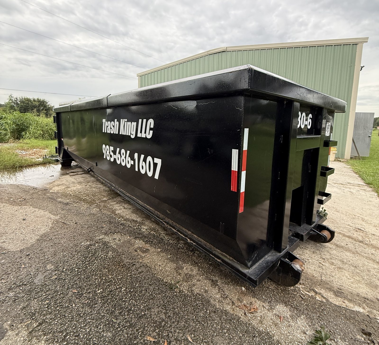A black 30-yard Trash King roll-off dumpster with Trash King LLC and 985-686-1607 on the side, set on a concrete pad beside a green metal building