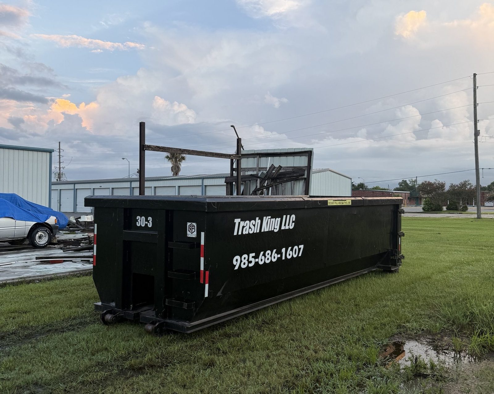 A black 30-yard Trash King LLC roll-off dumpster with 985-686-1607 on its side, set on a grass lot under a sunset sky